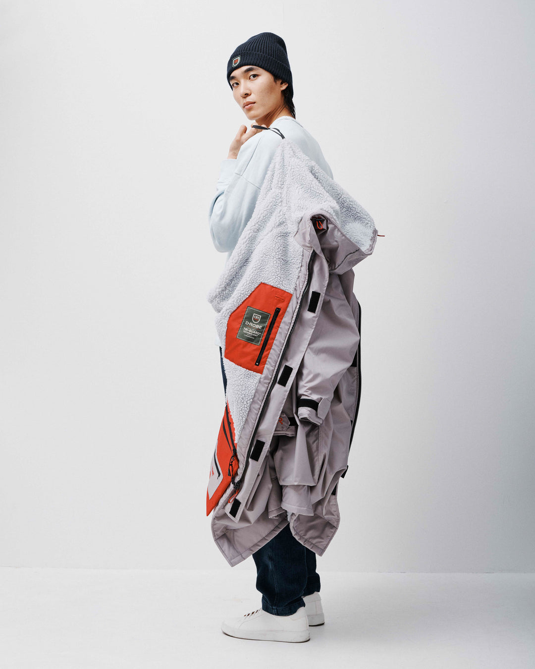 There is a man seated down wearing a grey dry-robe by D-Robe and black plants at Devon Beach. These hooded unisex changing robes are made from recycled materials, with a waterproof outer, fleece-lined inner, waterproof pockets, adjustable hood and cuffs. It is perfect for a stroll on the beach on a Spring Day.